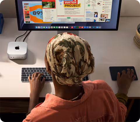 Masada oturan bir kişi Mac mini ile yazı yazıyor, Studio Display bağlı, Magic Keyboard, Magic Trackpad ve çeşitli eşyalar
