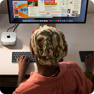 Masada oturan bir kişi Mac mini ile yazı yazıyor, Studio Display bağlı, Magic Keyboard, Magic Trackpad ve çeşitli eşyalar
