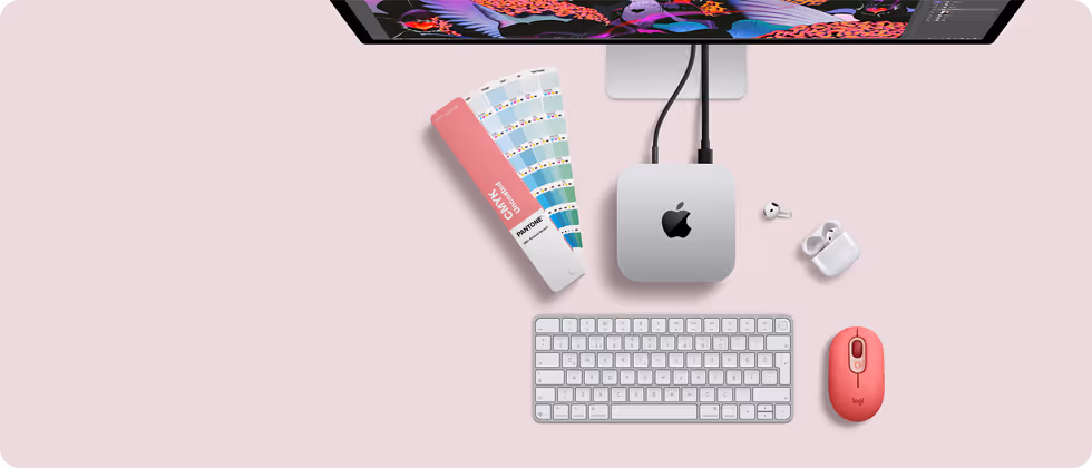 Mac mini, üstten görünüm, gümüş rengi alüminyum, yuvarlatılmış köşeler, ortalanmış parlak siyah Apple logosu, kabloyla bağlı, üstte Studio Display kısmen görünüyor, açık AirPod 4, Pantone renk kartları, sayısal tuş takımlı klavye ve mouse