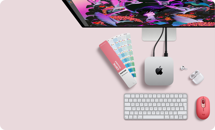 Mac mini, üstten görünüm, gümüş rengi alüminyum, yuvarlatılmış köşeler, ortalanmış parlak siyah Apple logosu, kabloyla bağlı, üstte Studio Display kısmen görünüyor, açık AirPod 4, Pantone renk kartları, sayısal tuş takımlı klavye ve mouse